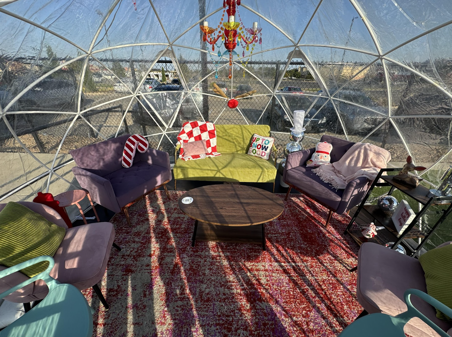 A cozy lounge inside a clear geodesic dome features colorful chairs, a green couch with decorative pillows, a red rug, and whimsical holiday decor, all bathed in sunlight from the transparent walls.